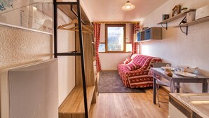 Een kleine kamer met een stapelbed, een tafeltje met een stoel, een radiator, een raam met gordijnen en een plank met spullen.