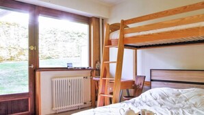 Een slaapkamer met een stapelbed, een bureau met een laptop, een radiator en een raam met uitzicht op groen.