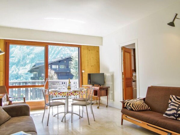 A living room with a sofa, a dining table with chairs, a TV, and a view of a mountain cabin through large windows.