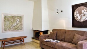 Een woonkamer met een bruine bank, een houten bankje, een open haard en twee kunstwerken aan de wand.