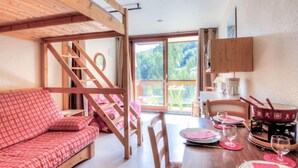 Une pièce chaleureuse avec un escalier en bois, un canapé à carreaux, une table à manger dressée pour deux personnes et une vue sur la verdure à l’extérieur.
