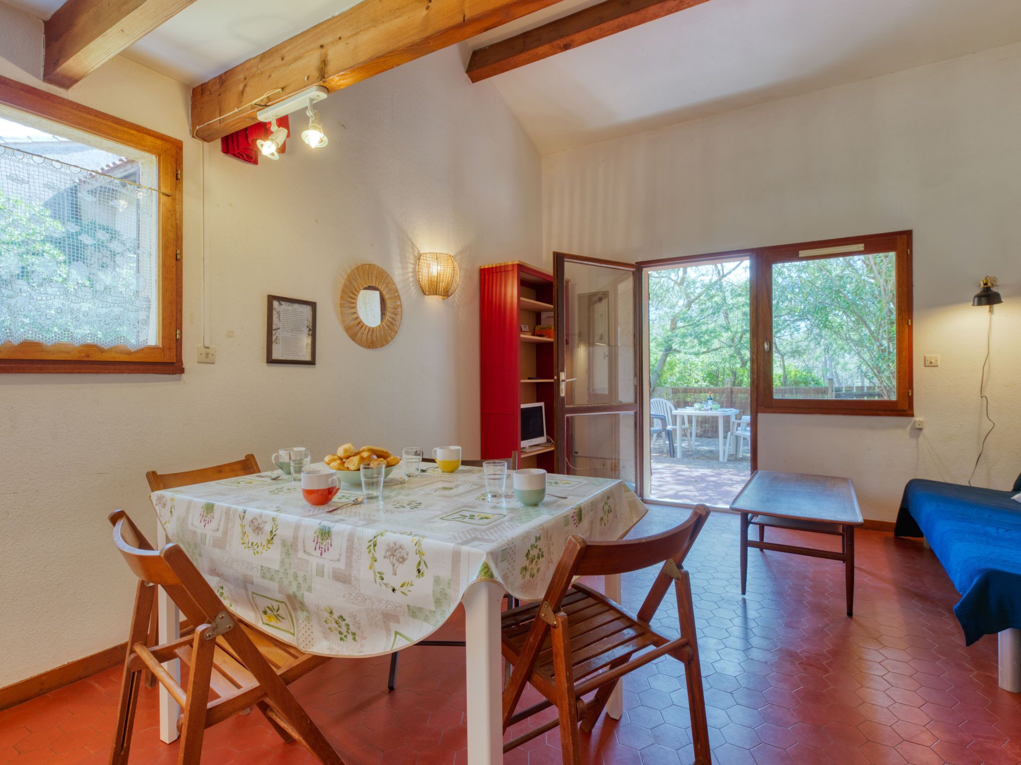 A dining area with a table set for four, wooden chairs, a red bookshelf, and a view of the outdoors through a glass door.