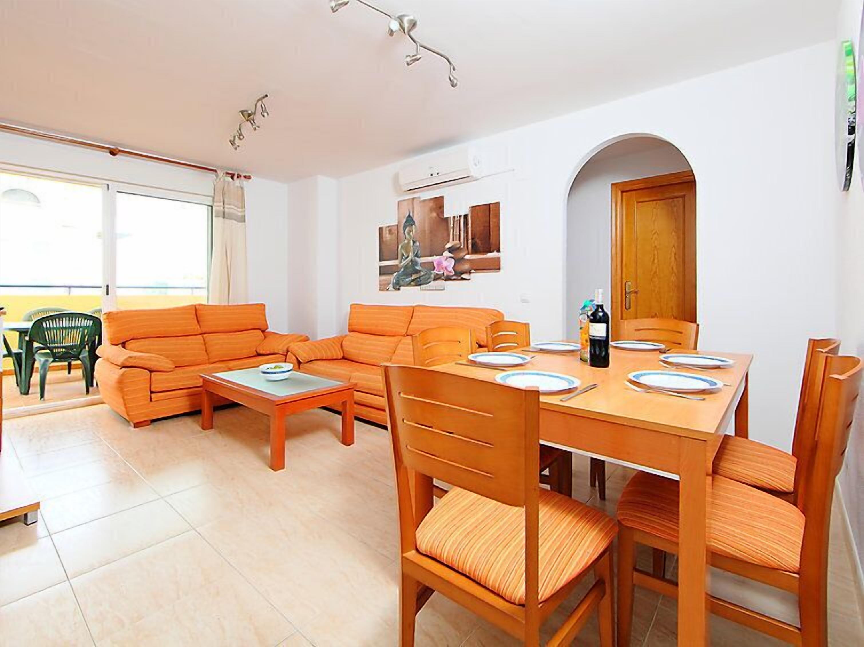 Un salon moderne comprenant un ensemble de canapés, une table à manger et des chaises. On y trouve une fenêtre avec des rideaux, une statue de Bouddha accrochée au mur et un appareil de climatisation.