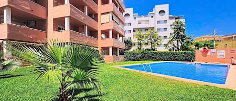 A swimming pool surrounded by well-maintained grass and palm trees, with multi-story buildings in the background.