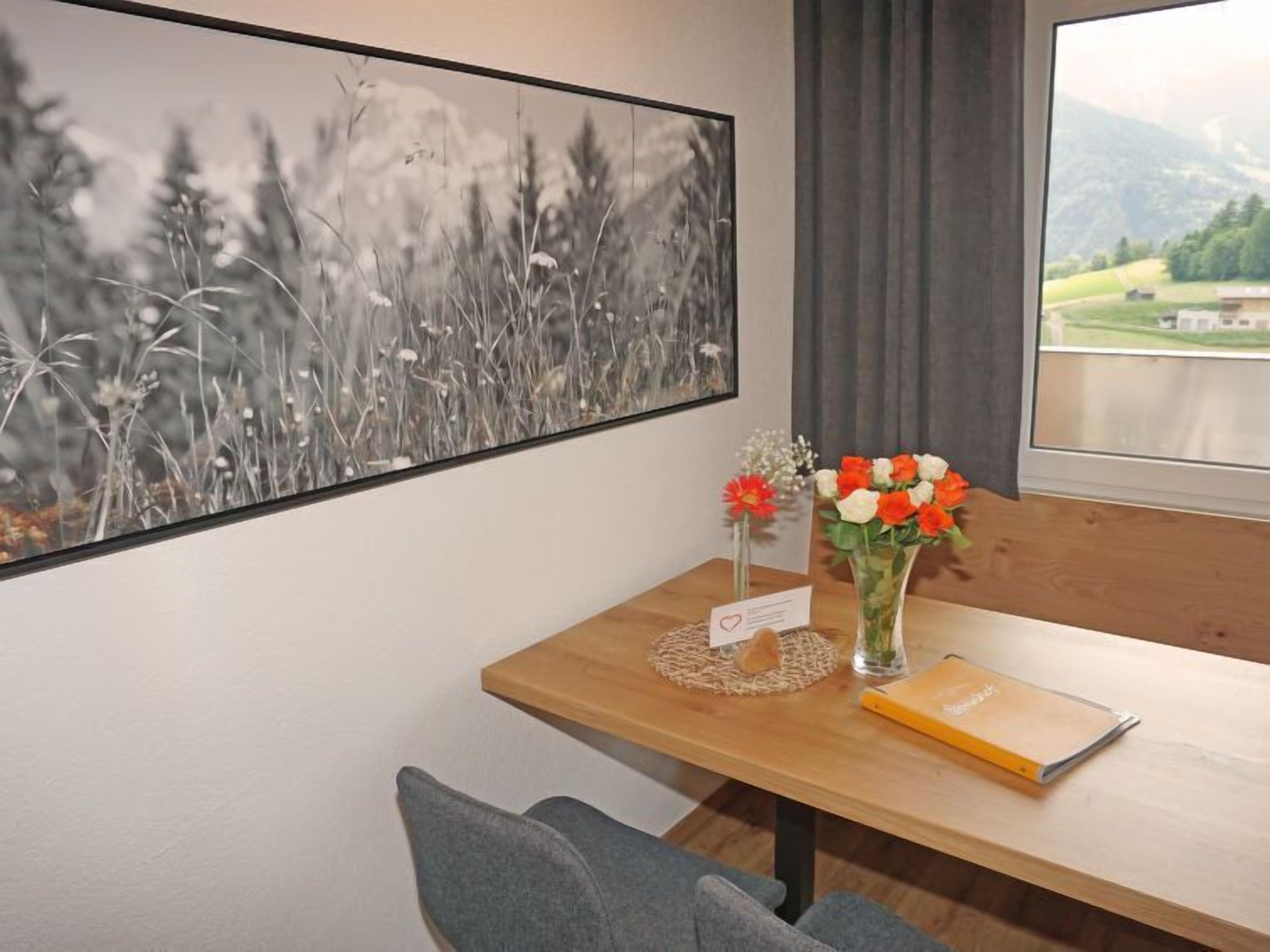 A room with a wooden table, a framed black and white landscape painting, a vase with red and white flowers, and a view of a green landscape through a window.