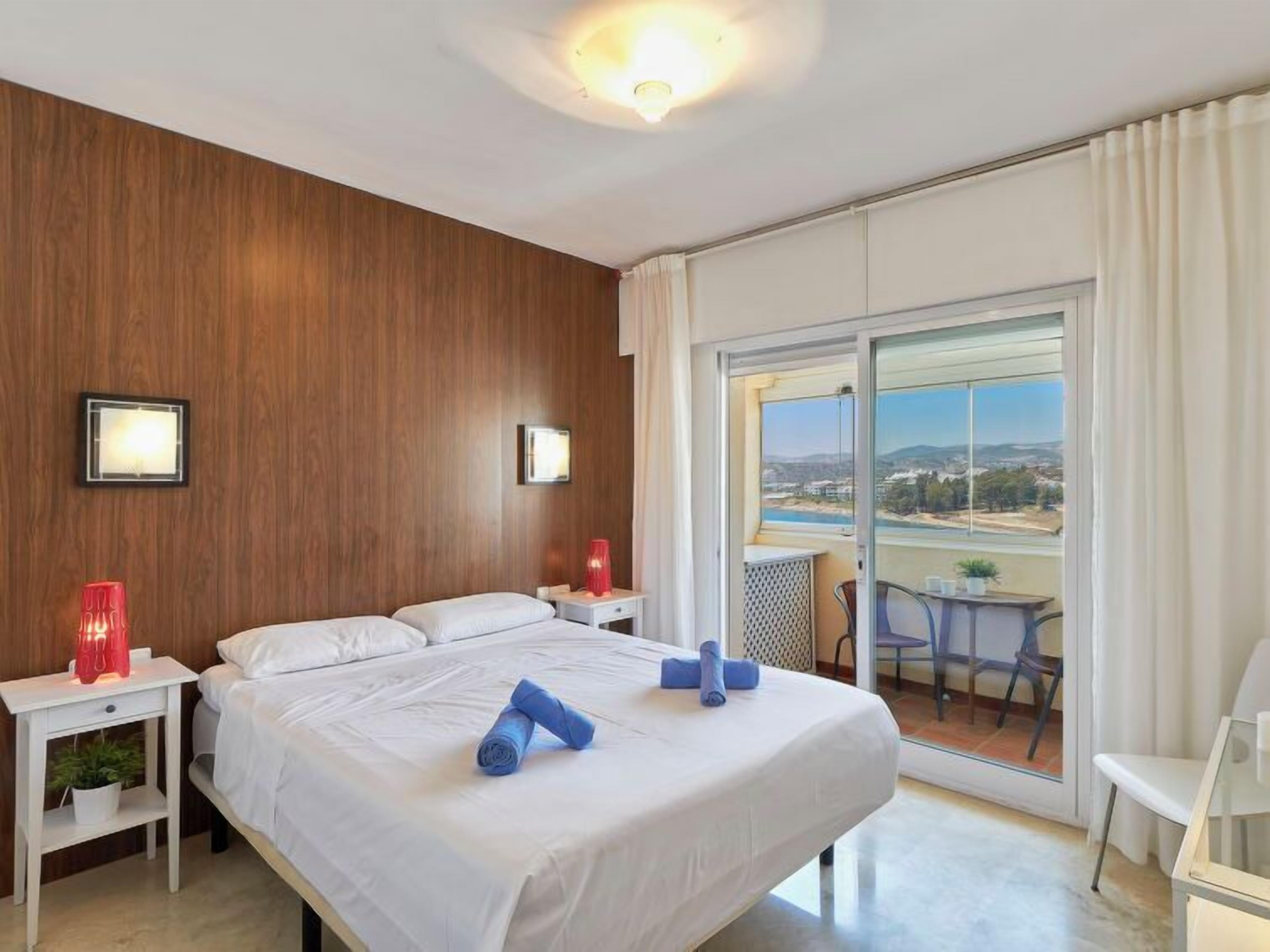 A hotel room with a large bed, two bedside tables, a red lamp, a small table with a chair, and a balcony with a view.