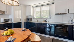 Una cocina moderna con armarios oscuros, una mesa de comedor de madera con frutas, una cafetera y una ventana que deja entrar luz natural.