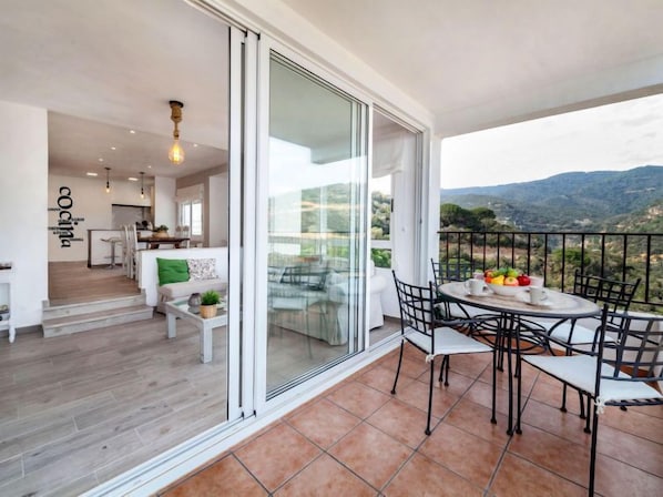 Ein Balkon mit Tisch und Stühlen, eine Schiebetür zu einem Wohnbereich und Blick auf die Landschaft.