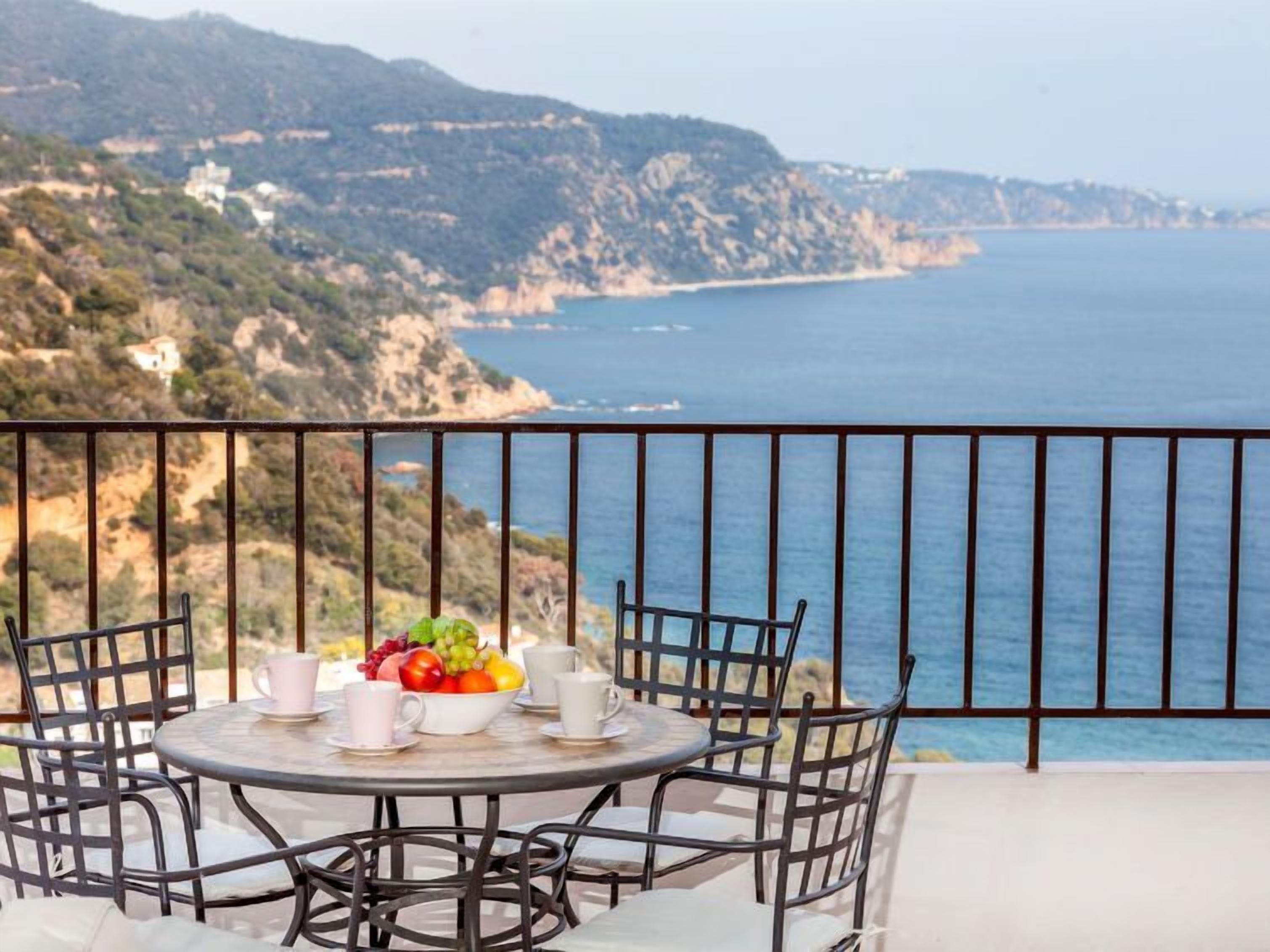 A round table with four chairs and a fruit bowl on a balcony overlooking a coastal landscape.
