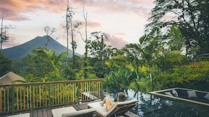 Una terraza de madera con sillones de descanso y piscina, rodeada de exuberante vegetación tropical y una montaña al fondo.