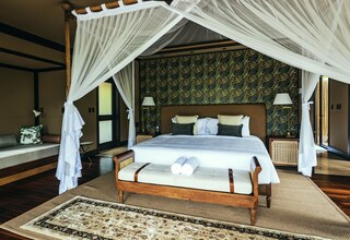 A four-poster canopy bed with a wooden frame and white bedding, a patterned wall behind, and a view of the outdoors through a glass door.