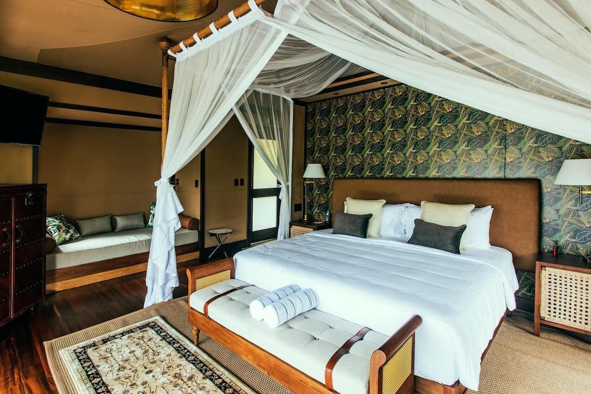 A four-poster canopy bed with a headboard, a bedside table with a lamp, a wooden wardrobe, and a patterned wall.