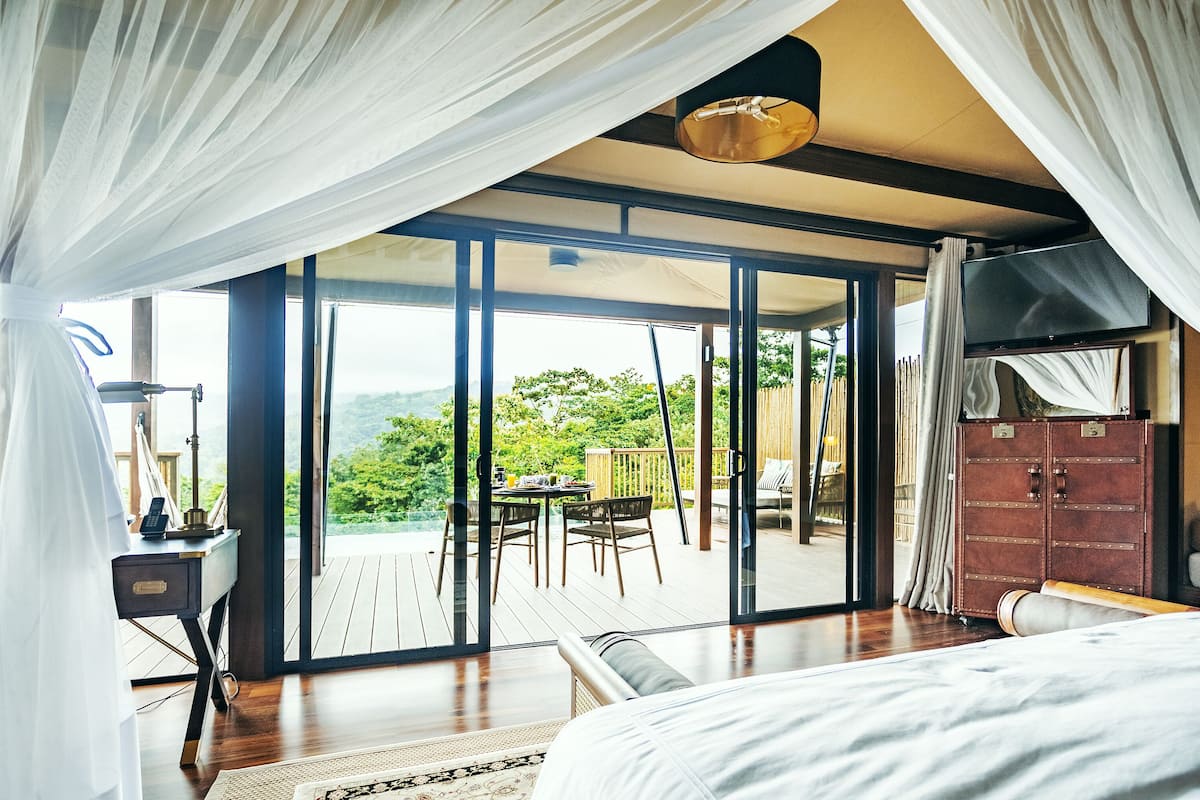 A bedroom with a large bed, a desk with a lamp, and a view of the outdoors through a sliding glass door.