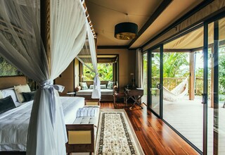 A bedroom with a canopy bed, a sitting area with a sofa and chairs, and a view of a hammock outside.
