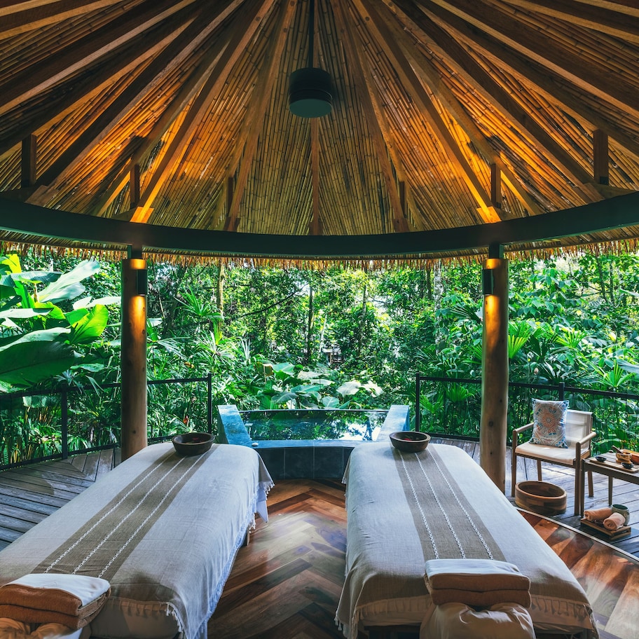 A room with two beds, a small table, and chairs, surrounded by lush greenery.