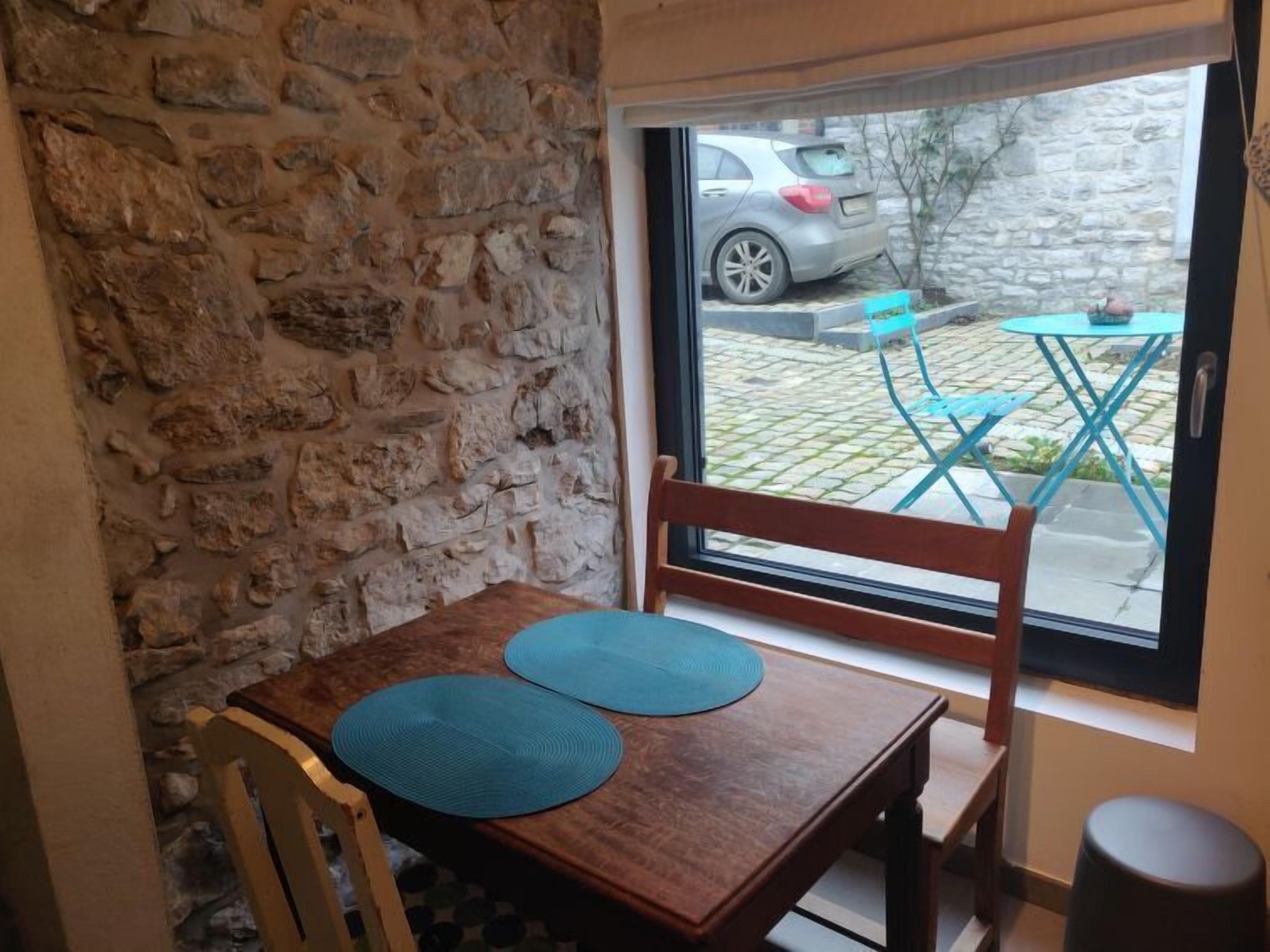A corner of a room with a stone wall, a wooden table with two blue placemats, a wooden chair, and a view of a car and outdoor seating area through a glass door.