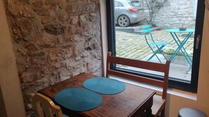 Een hoek van een kamer met een stenen muur, een houten tafel met twee blauwe placemats, een houten stoel en zicht op een auto en een terras door een glazen deur.