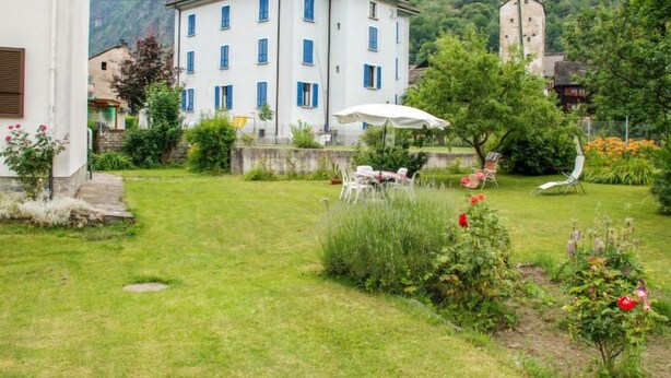 Un edificio bianco con persiane blu, un giardino con un ombrellone bianco e montagne sullo sfondo.