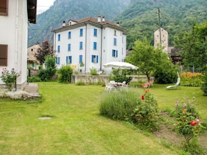 A white building with blue shutters, a garden with a white umbrella, and a mountain backdrop.