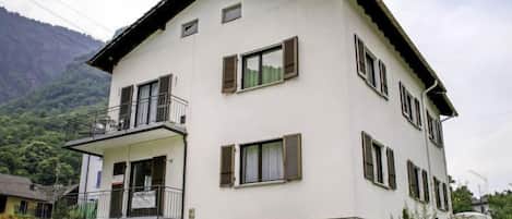 A two-story building with a balcony on the second floor, stone foundation, and a mountainous backdrop.