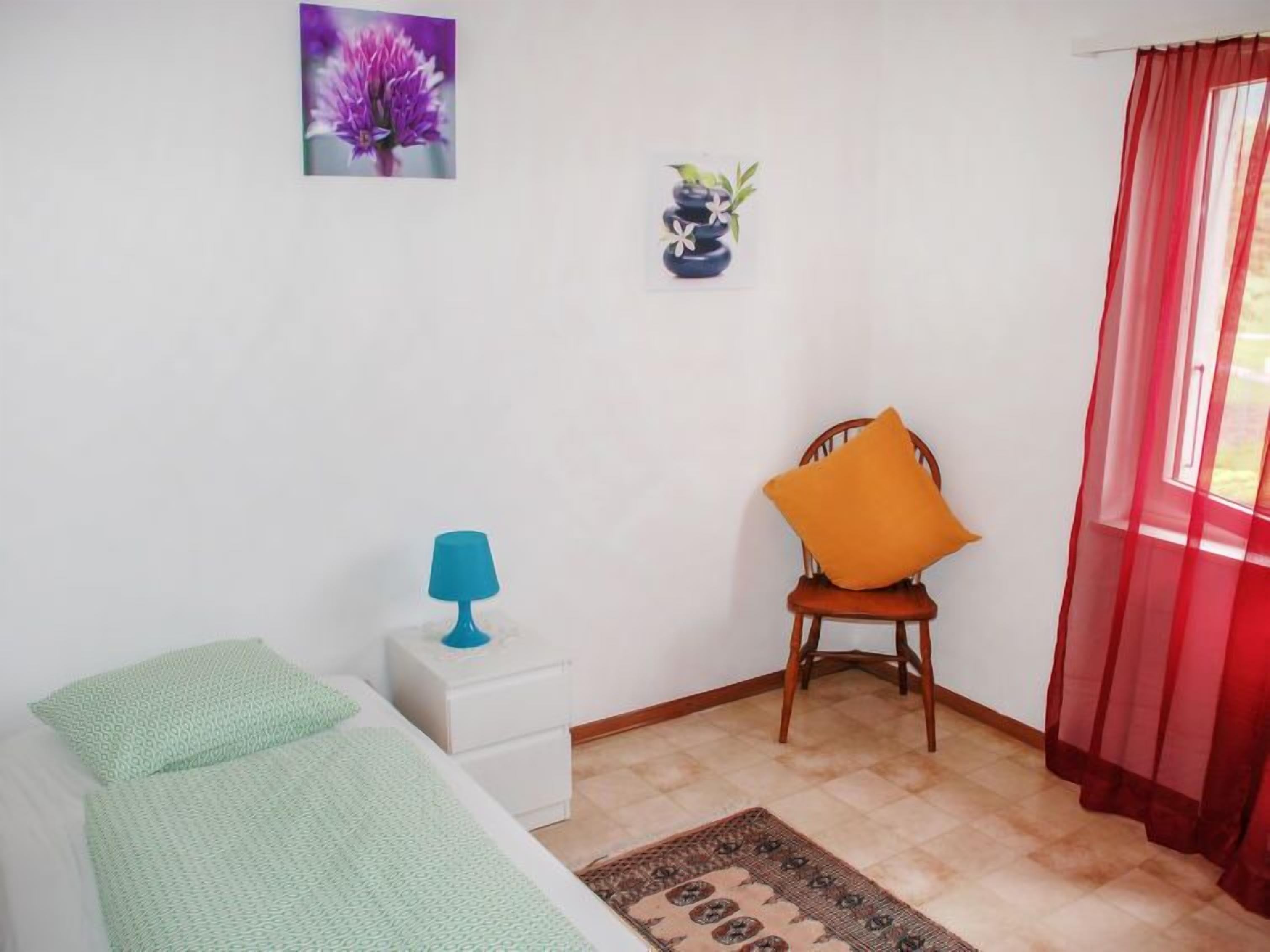 Quarto com cama, mesa de cabeceira, cadeira com almofada laranja, pequena mesa com abajur azul, vaso com flores e janela com cortinas vermelhas.