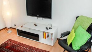 Un soggiorno con una TV a parete, un mobile bianco per l'intrattenimento, una sedia da ufficio nera con cuscini verdi e gialli, un tappeto rosso con motivo e una lampada da terra.