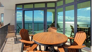 Een balkon met een houten tafel en stoelen, grote ramen en uitzicht op de oceaan.