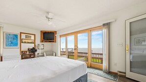A hotel room with a large bed, a ceiling fan, a TV mounted on the wall, a bookshelf, and a view of the ocean through a sliding glass door.