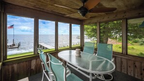 A wooden deck with a table and chairs, overlooking a body of water with a dock and an American flag.