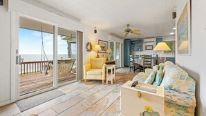 A spacious living area with a large sliding glass door leading to a balcony, a ceiling fan, a yellow armchair, a patterned sofa, and a kitchen area with blue cabinets.