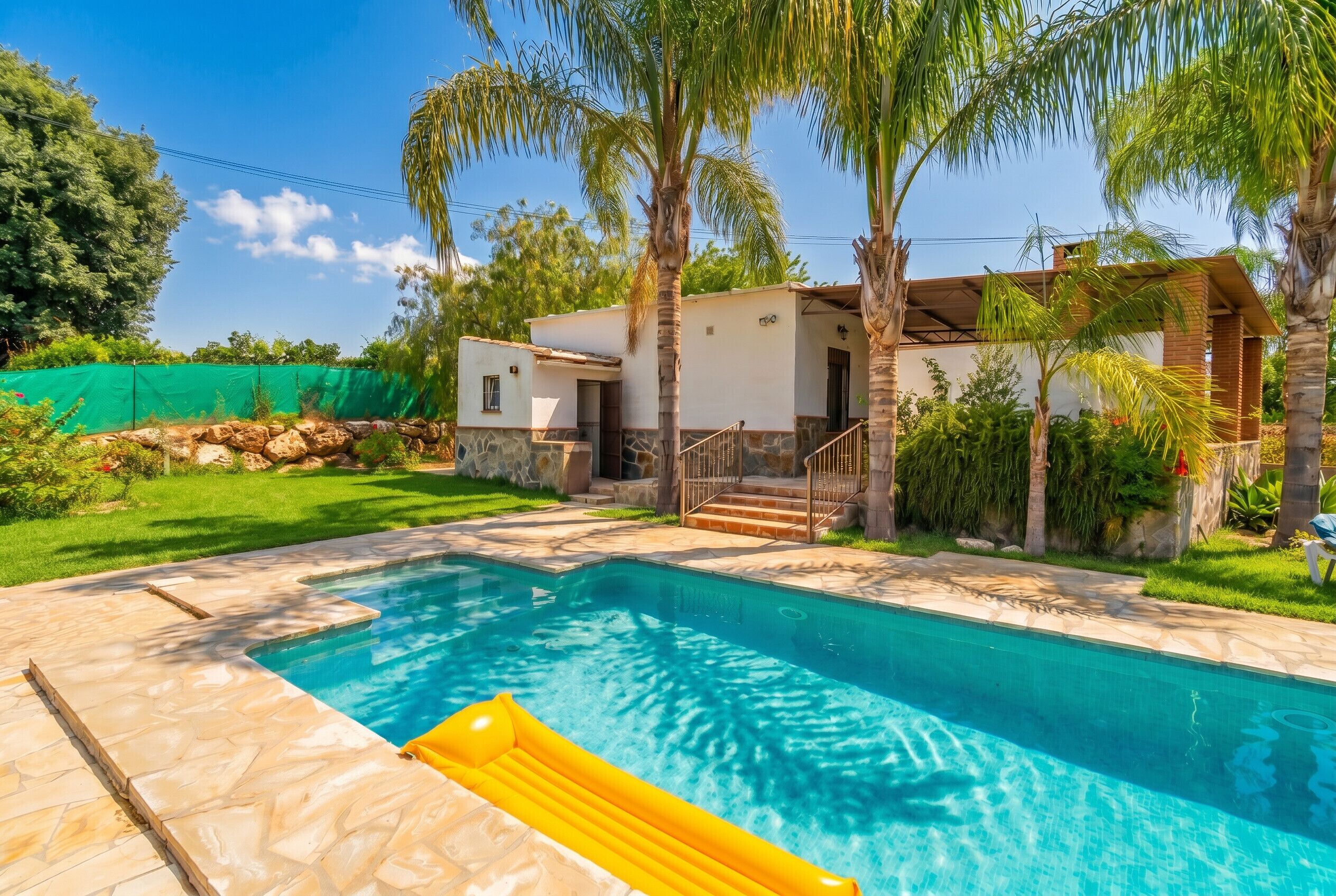 Uma piscina com um boi de água amarelo, uma casa com fachada de pedra e um jardim bem cuidado com palmeiras.