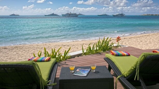 A beachside lounge with two green cushioned chairs, a table with a magazine and glasses of orange juice, and a view of the ocean with several boats. - Vittoria (6 bedrooms) - Beachfront villa, Simpson Bay, St. Maarten (Simpson Bay)