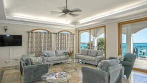 A spacious living room with grey sofas, a glass coffee table, and a ceiling fan. Large windows offer a view of the sea. - Vittoria (6 bedrooms) - Beachfront villa, Simpson Bay, St. Maarten (Simpson Bay)