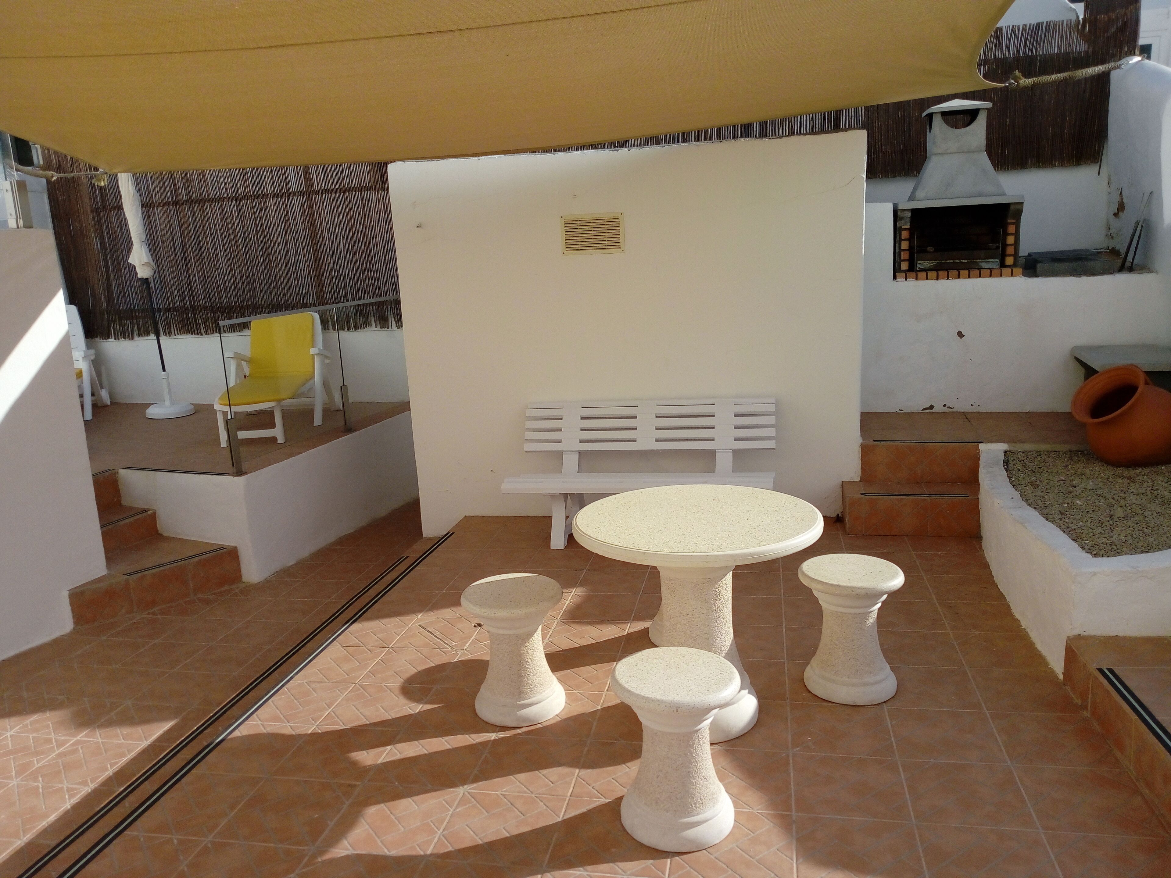 A patio area with a white table and two stools, a yellow chair, a white bench, and a small outdoor oven.