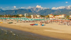 A beach with umbrellas and boats, buildings in the background, and mountains.