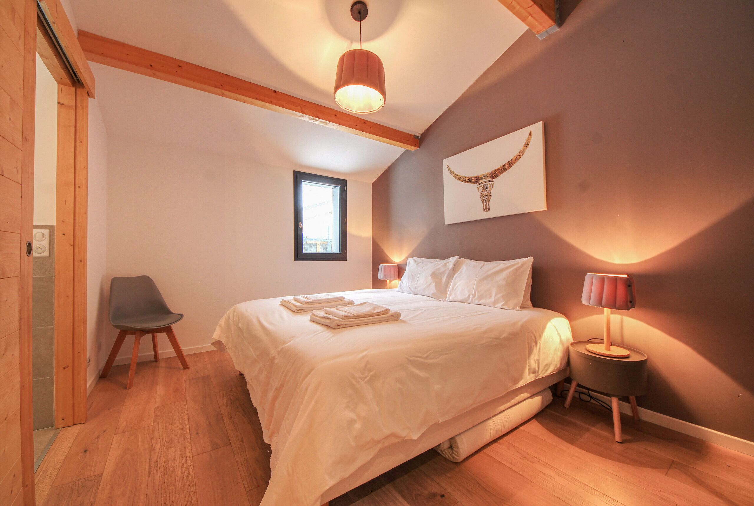 Un dormitorio con cama, lámparas de noche, una silla y una ventana.