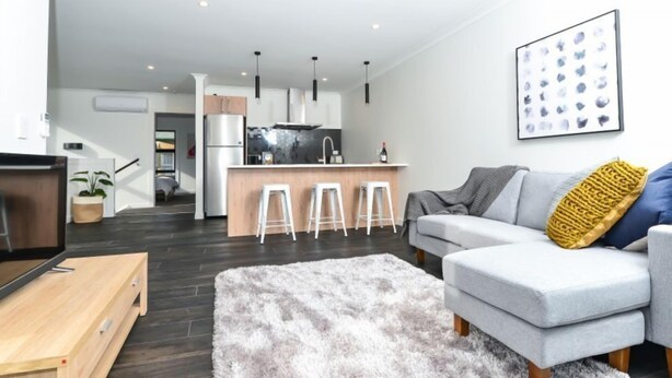 A modern living room with a grey sofa, a fluffy rug, and a kitchen in the background. - Central Park Gemini Apartments (Hamilton)