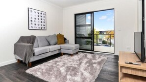 A modern living room with a grey sectional sofa, a fluffy rug, a flat-screen TV, and a large sliding glass door leading to a balcony. - Central Park Gemini Apartments (Hamilton)