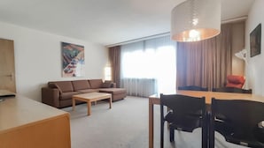 Ein modernes Wohnzimmer mit Sofa, Couchtisch, Esstisch und Stühlen. An der Wand hängt ein Gemälde, ein Fenster mit Vorhängen und eine Hängelampe.