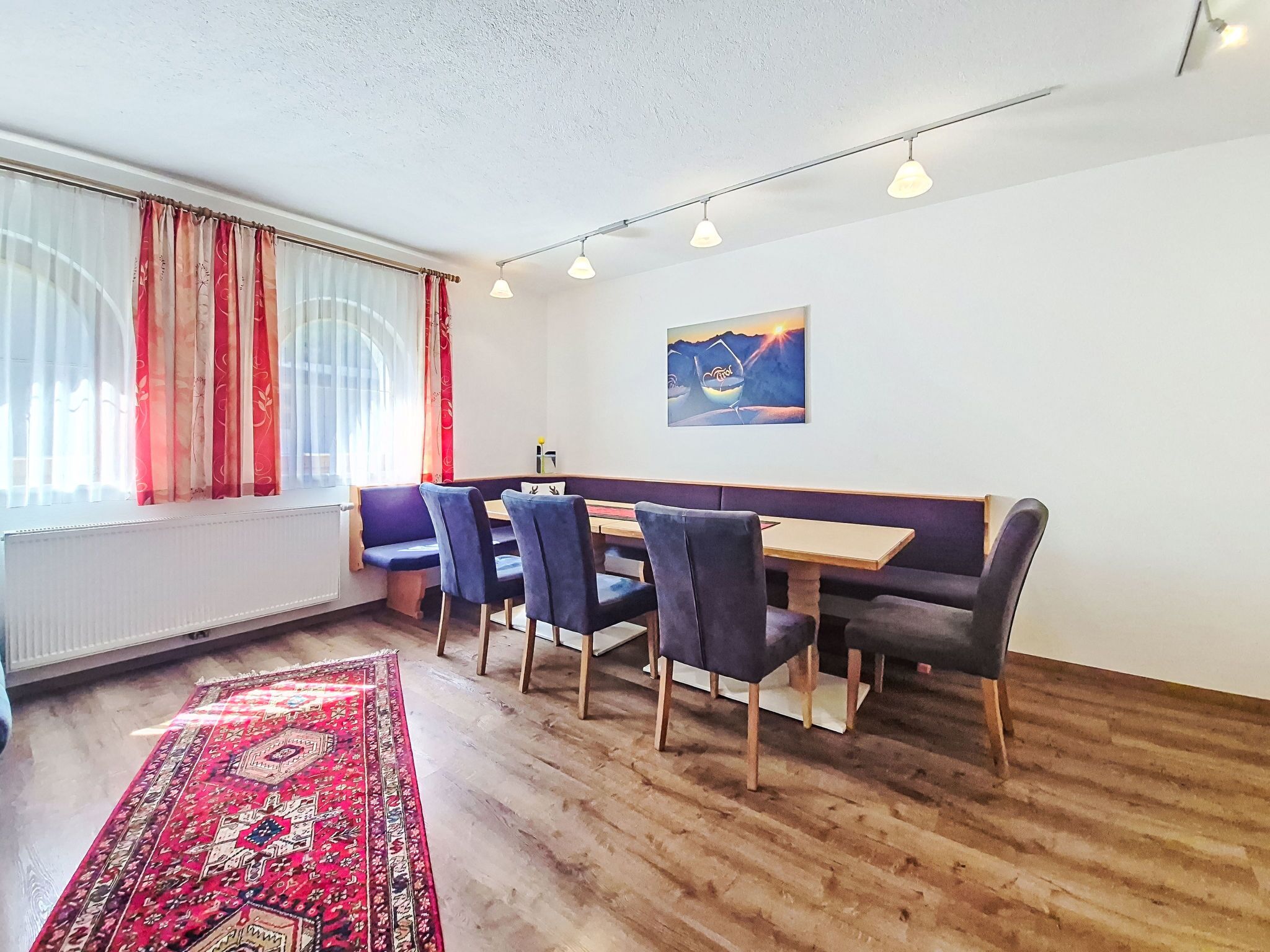 Een kamer met een houten vloer, een rood tapijt met patroon, een bankje en een tafel met stoelen.