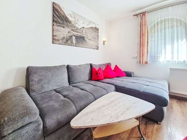 A grey sectional sofa with red cushions, a wooden coffee table, a framed picture on the wall, and a window with sheer curtains.