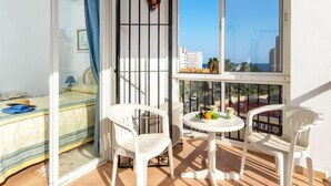 Un balcon équipé d’un lit, de deux chaises, d’une table avec un panier de fruits, et offrant une vue sur les bâtiments et les palmiers.