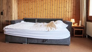 Un dormitorio con pared de paneles de madera, una cama con ropa de cama gris, una mesita de noche con una lámpara y una ventana con cortinas.