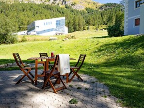 Ein Holztisch mit Stühlen steht auf einer gepflasterten Terrasse mit Blick auf ein modernes Gebäude und bewaldete Hügel im Hintergrund.