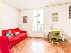 A bright living room with a red sofa, a dining table with chairs, and a herringbone wooden floor.