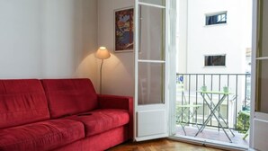En rød sofa i et rum med trægulv, en hvid dør der fører til en balkon med et bord og stole, samt en lampe.