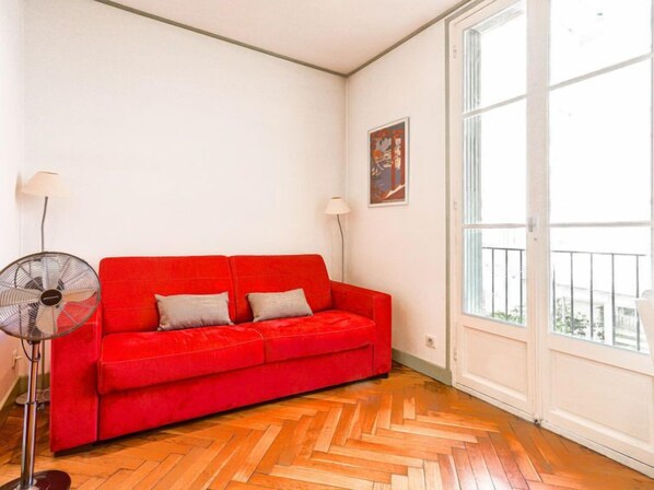 A red sofa in a room with a herringbone floor, a standing fan, and a large window.