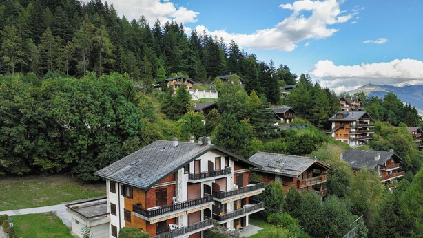 Ein mehrstöckiges Gebäude mit Balkonen, umgeben von üppiger Vegetation und Bergen im Hintergrund.