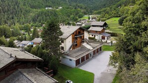 Ein Bergdorf mit traditionellen Holzhäusern, ein zentrales Gebäude mit Balkon und Blick auf den umliegenden Wald und die Berge.