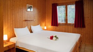 Ein Zimmer mit Holzwänden, einem großen Bett, zwei Nachttischen mit Lampen, einem Fenster mit roten Vorhängen und einem kleinen gerahmten Bild an der Wand.